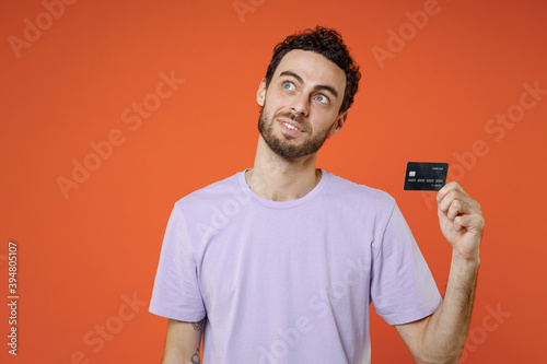 Smiling pensive young bearded man 20s wearing casual basic violet t-shirt standing holding in hand credit bank card looking aside up isolated on bright orange color wall background studio portrait. © ViDi Studio