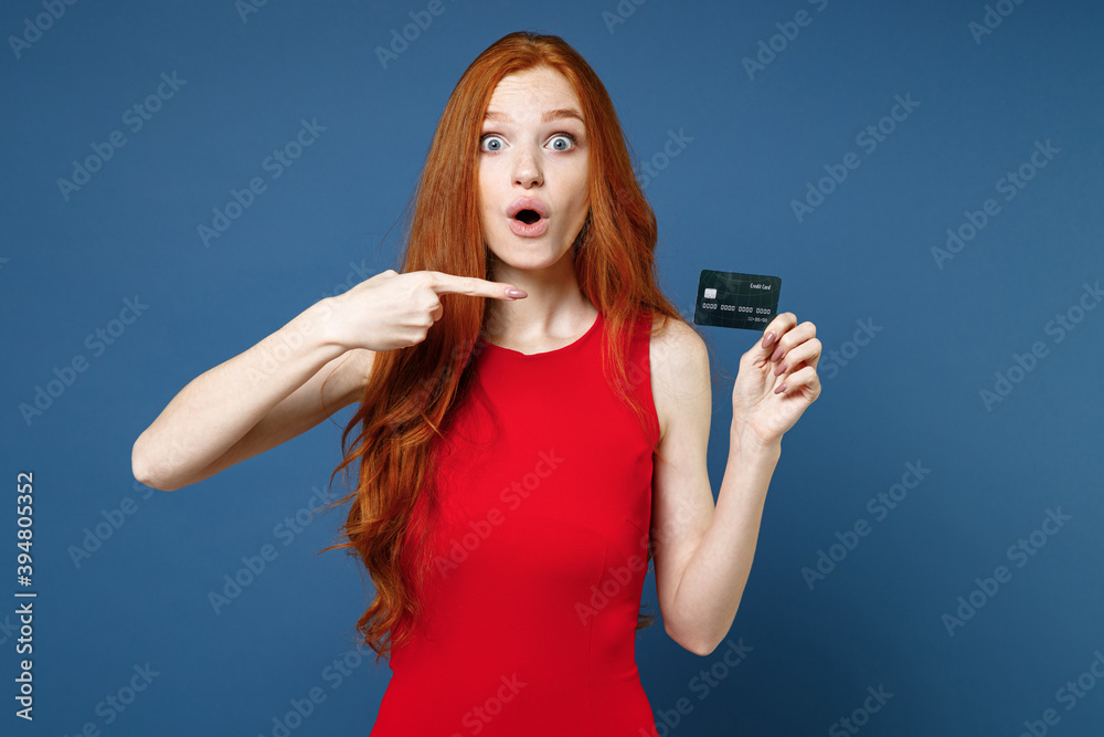 Shocked amazed young redhead woman 20s wearing bright red elegant evening dress standing pointing index finger on credit bank card looking camera isolated on blue color background studio portrait. Shocked amazed young redhead woman 20s wearing bright red elegant evening dress standing pointing index finger on credit bank card looking camera isolated on blue color background studio portrait.