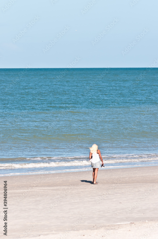 back view, very far distance of a senior female with long blond hair, walking a tropical shoreline of a sandy beach on gulf of Mexico on a sunny day back view, very far distance of a senior female with long blond hair, walking a tropical shoreline of a sandy beach on gulf of Mexico on a sunny day
