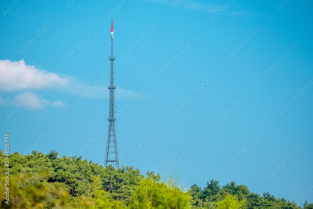 Telecommunication tower with signal amplifiers for mobile communications and antennas is installed in a rural area on a grassy field with trees.
