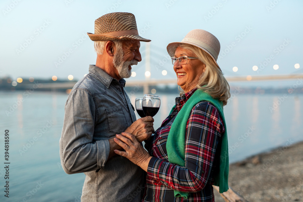 A happy elderly couple is drinking wine on the riverside. 