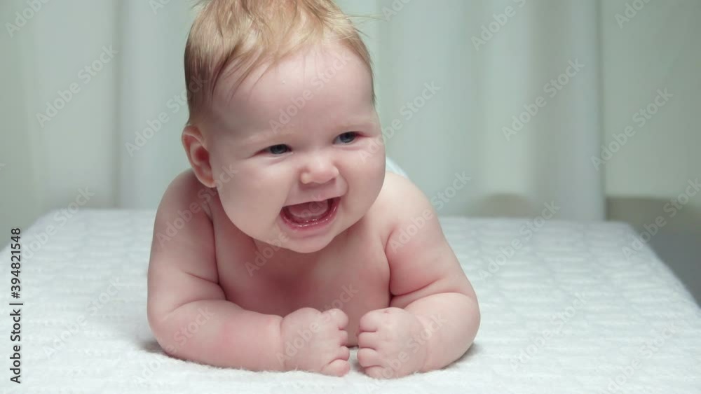 The little cute baby is laying on the stomach on a white blanket and laughing happily The little cute baby is laying on the stomach on a white blanket and laughing happily