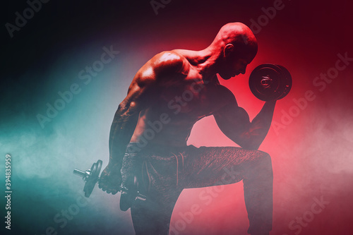 Brutal strong athletic Bodybuilder posing with dumbbells. Double light and smoke effect on athlete. Body building and healty life concept