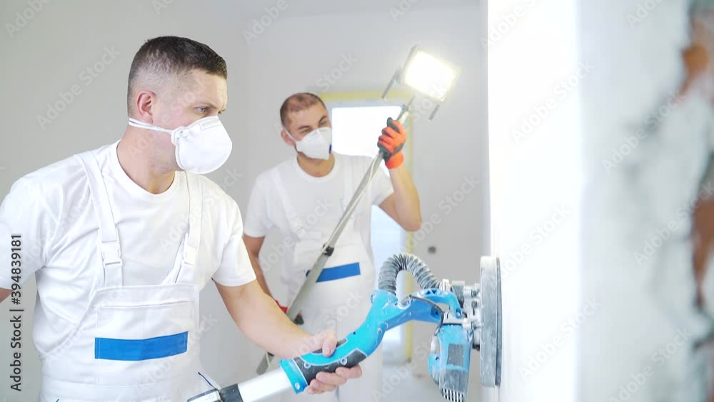 Plasterer smoothing wall surface with wall machine sandpaper grinding machine. Two master builders sanding a white plaster wall. man in overalls grinds the surface in respirator. experienced repairman