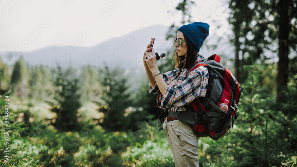 Stylish woman traveler in a hat stands on the top of the mountain. Hipster girl on the top of the mountain, resting and taking pictures on the phone. Atmospheric moment. Wanderlust and travel concept