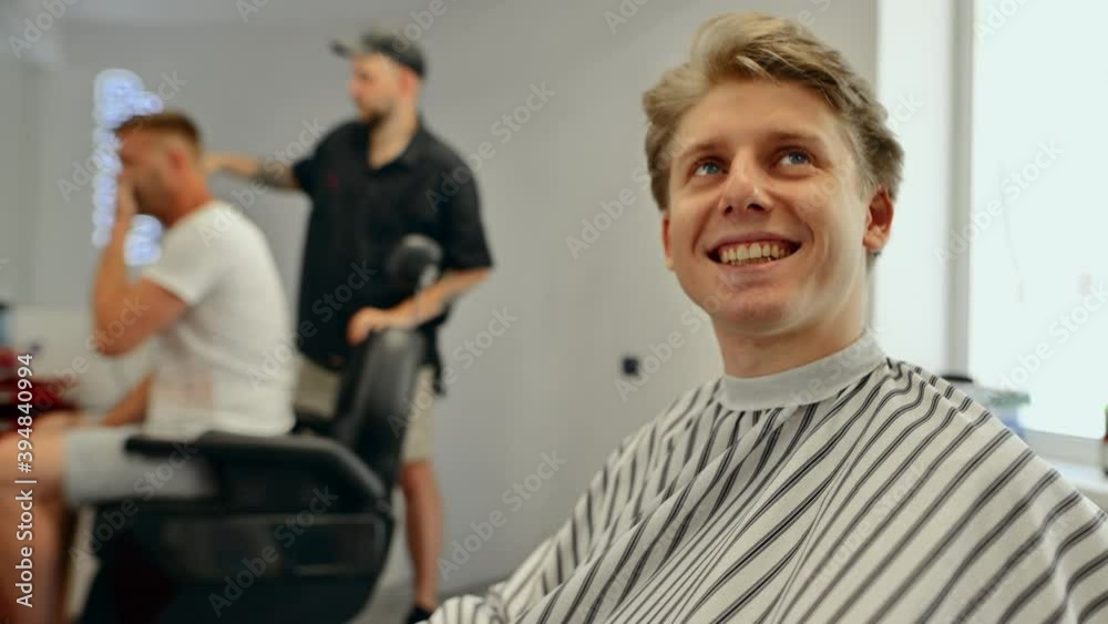 Positive young male client sitting in a peignoir in a barbershop chair, looking at the camera with a smile on his face and showing his fingers gesture OK Positive young male client sitting in a peignoir in a barbershop chair, looking at the camera with a smile on his face and showing his fingers gesture OK
