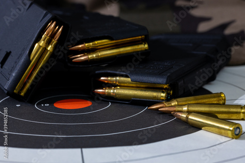Loaded rifle magazines with loose rounds of ammunition scattered on top of a paper target with a camouflage background. Background and foreground blurred with a shallow depth of focus.
