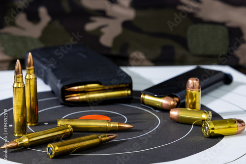 Loaded rifle and pistol magazines with loose rounds of ammunition scattered on top of a paper target with a camouflage background. Background and foreground blurred with a shallow depth of focus.