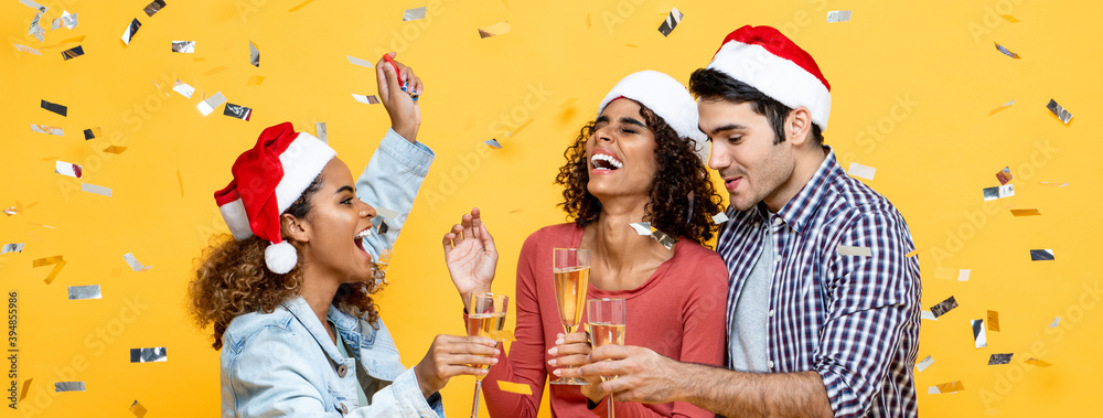 Group of three happy diverse friends celebrating Christmas drinking champagne on yellow studio bannrer background