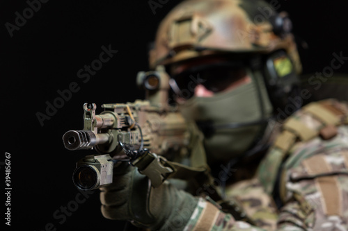 Close up of a soldier in camouflage and full military gear, wearing a helmet, glasses and protective mask, raises a rifle to his face and taking aim, preparing to shoot, isolated on black background © satyrenko
