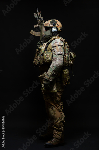 Full length side view portrait of an army soldier standing in full military uniform, wearing a bulletproof vest, helmet, glasses and mask, holding up a submachine gun isolated on a black background