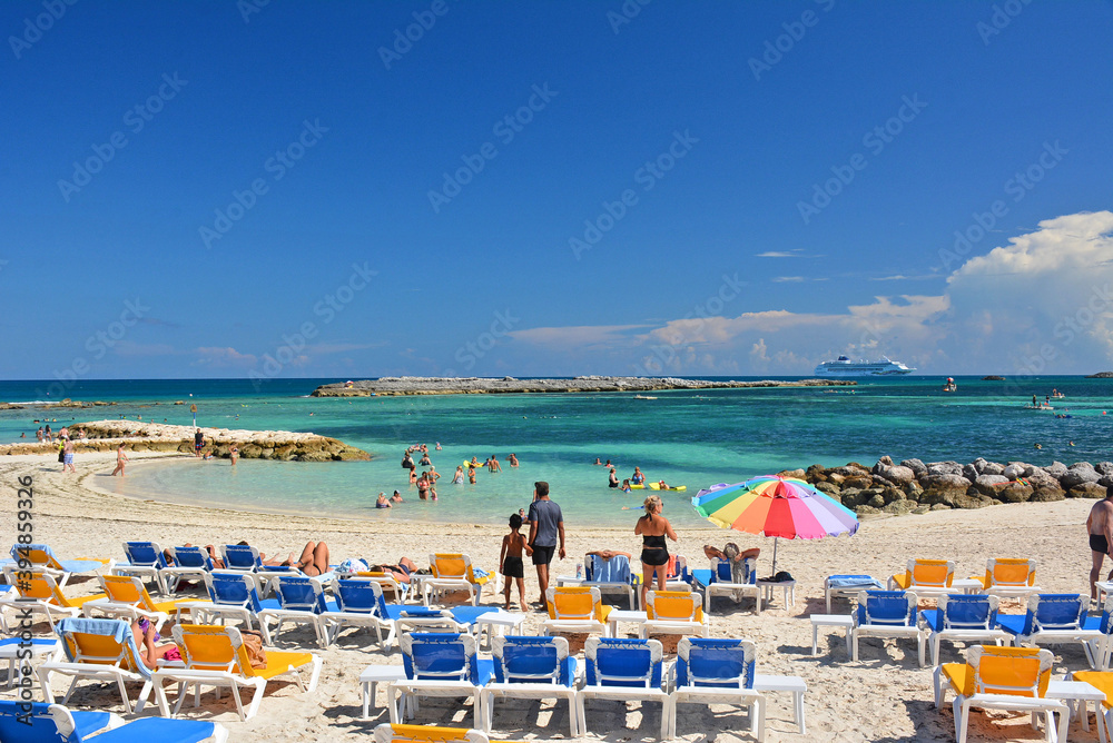 Tourists with beach chairs and umbrellas at a private island beach in the Bahamas  with cruise ship in the background