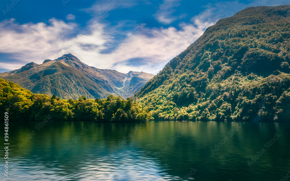 Sunlit mountain range covered in rainforest and reflections in the fjord's water on a beautiful summer day at Doubtful Sound in New Zealand, South Island.