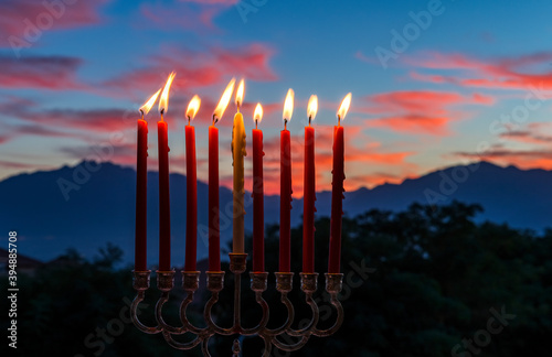 Glitter lights of candles on menorah are traditional symbols for Jewish Hanukkah Holiday of Light. Selective focus on candles. Background with blurred dramatic morning sky and mountains as background