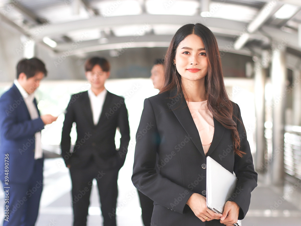confident beautiful Asian business woman in black suit holding computer laptop, standing in front of her male teamwork, smiling and looking at camera. confident beautiful Asian business woman in black suit holding computer laptop, standing in front of her male teamwork, smiling and looking at camera.