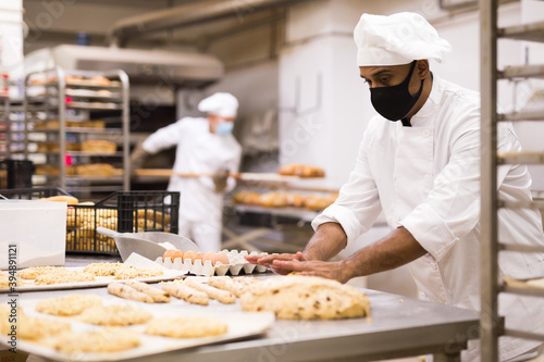 Fotografiet male baker in mask and white uniform rolling out dough in kitchen