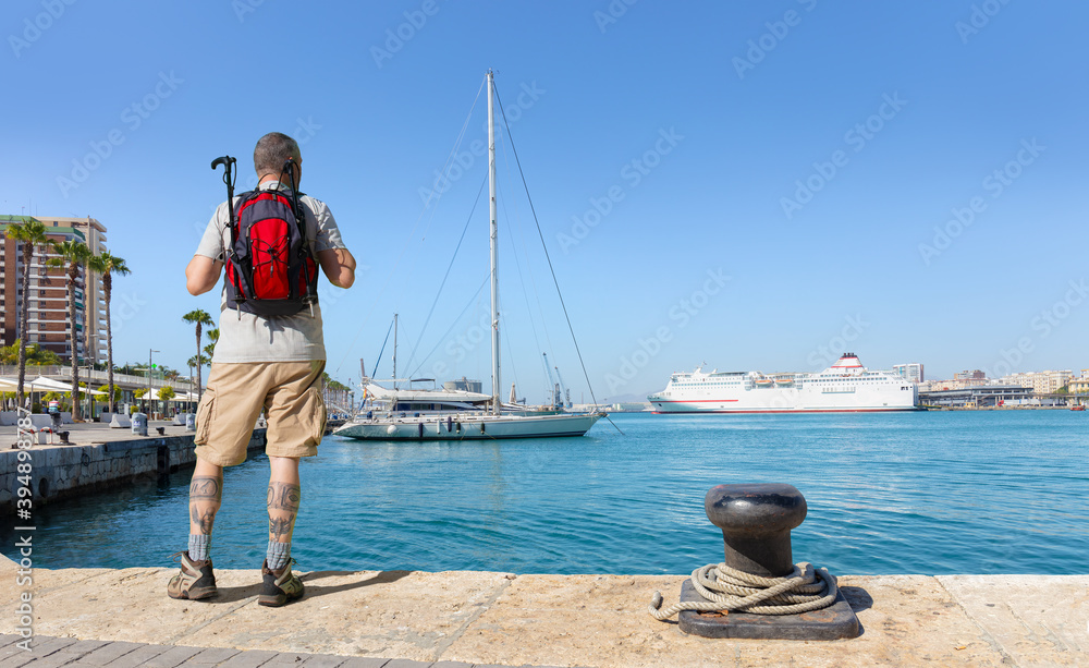 A male hiker with a backpack looks out over the port of Malaga in Andalusia. It's a summer day with blue skies. There are sailboats and a white ferry in the background.