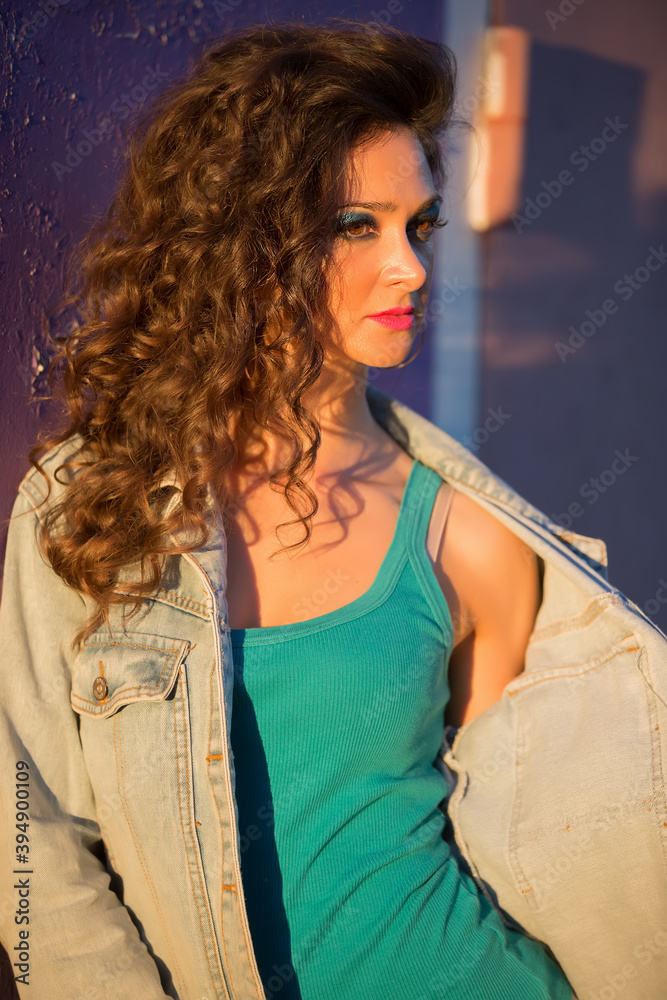 Beautiful young woman in a denim jacket on the background of a peeled purple wall. Girl with bright makeup in the style of the eighties on the street. Beautiful young woman in a denim jacket on the background of a peeled purple wall. Girl with bright makeup in the style of the eighties on the street.
