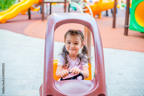 Cheerful little girl with two ponytails sits in a children's car on the playground in the park