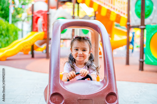 Cheerful little girl with two ponytails sits in a children's car on the playground in the park