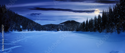 snow covered mountain lake at night. green spruce trees on the shore in full moon light. clouds on the sky