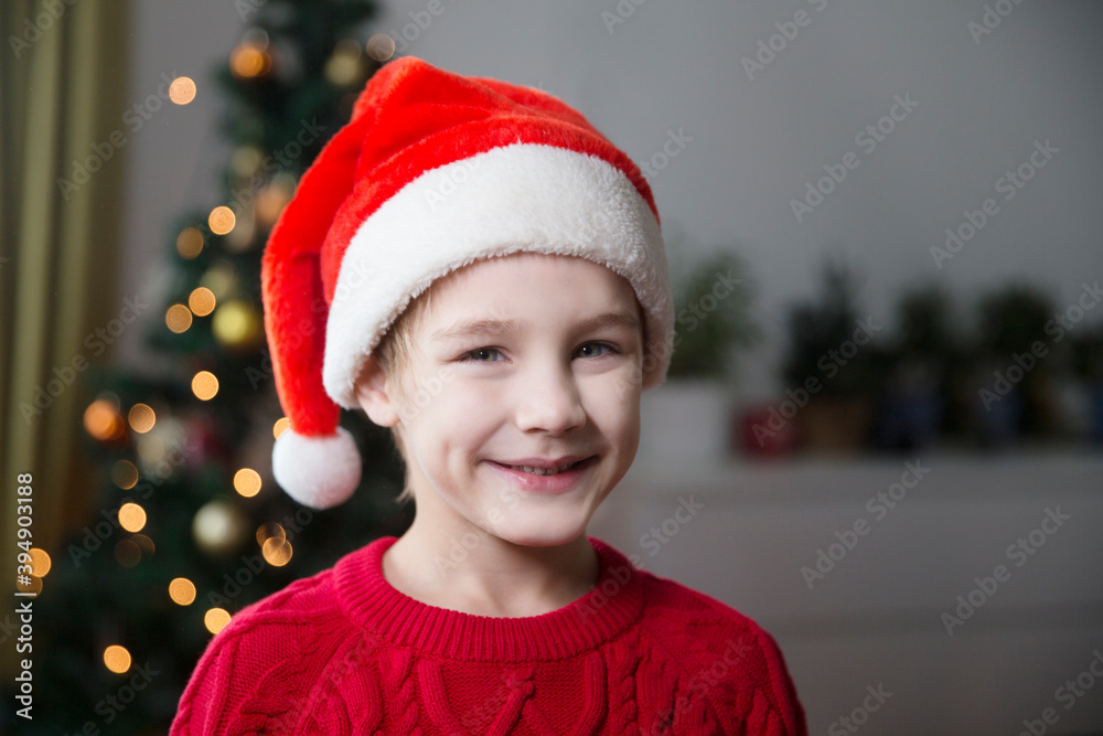 smiling cheerful Kid boy in a Santa hat and a red sweater celebrates Christmas at home.