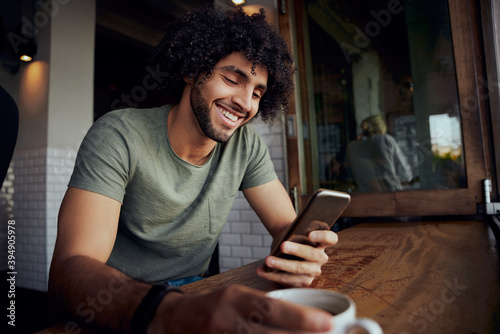 Handsome smiling young man sitting in cafe holding coffee mug while using smartphone