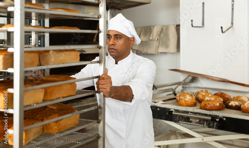 Focused male chef in uniform pushing rack trolley with fresh bread in bakery kitchen
