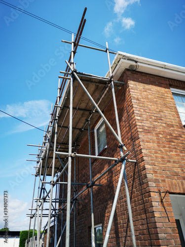 Traditional red brick detached house undergoing renovation or repairs with scaffolding and boards attached to the house. Low angle, corner view with blue sky.