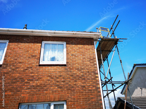 Traditional red brick detached house undergoing renovation or repairs with scaffolding and boards attached to the house. Low angle, corner view with blue sky.