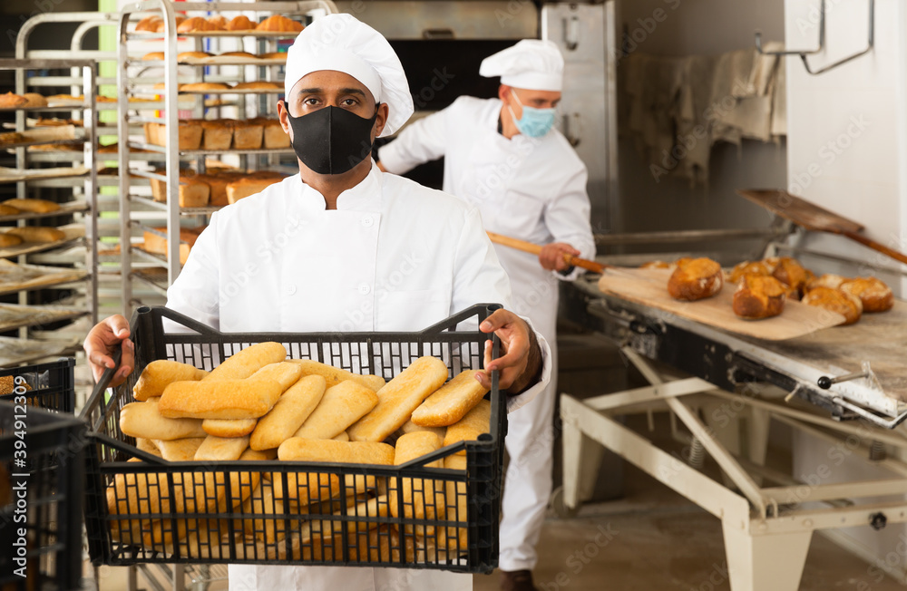 Focused young man in face mask for viral protection working in small bakery, carrying fresh baked bread in crate