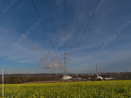 Bottom view of a power line with pylon connecting to nuclear power plant in Neckarwestheim, Germany with yellow blooming rapeseed flowers below on sunny day in autumn season with few clouds. photo