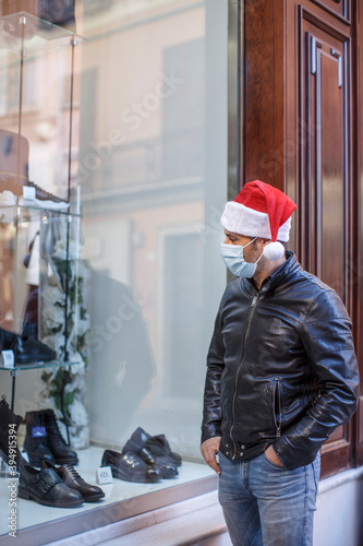 Uomo con cappello da babbo natale, giacca in pelle e mascherina chirurgica, gira tra le vetrinedi un negozio in centro 