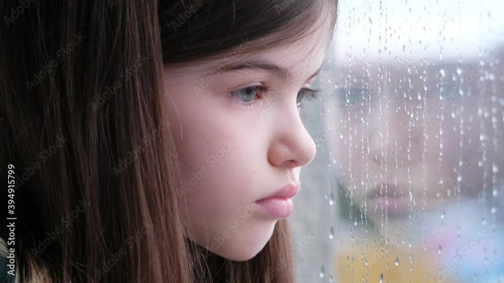 A sad alone little / young girl sits and looks out the window. It is raining outside the window, drops are running down the glass. Isolation, coronavirus,  concept
