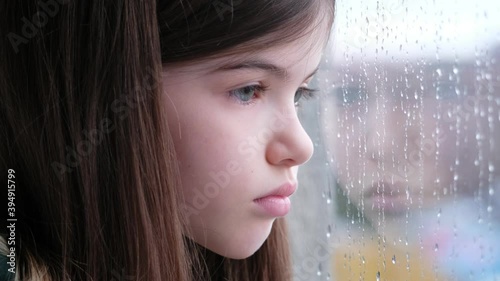 A sad alone little / young girl sits and looks out the window. It is raining outside the window, drops are running down the glass. Isolation, coronavirus,  concept