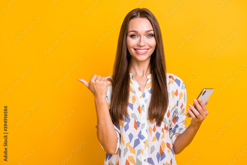 Photo portrait of girl pointing thumb at blank space holding phone in one hand isolated on vivid yellow colored background Photo portrait of girl pointing thumb at blank space holding phone in one hand isolated on vivid yellow colored background