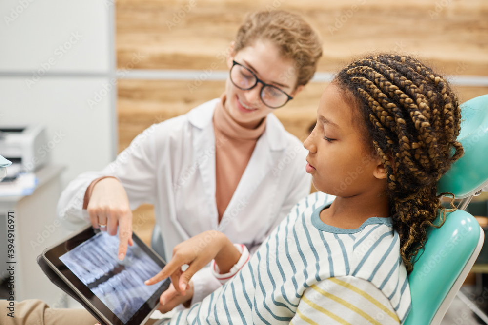 Little girl sitting on chair and looking at x-ray image of her teeth together with dentist at dental clinic Little girl sitting on chair and looking at x-ray image of her teeth together with dentist at dental clinic