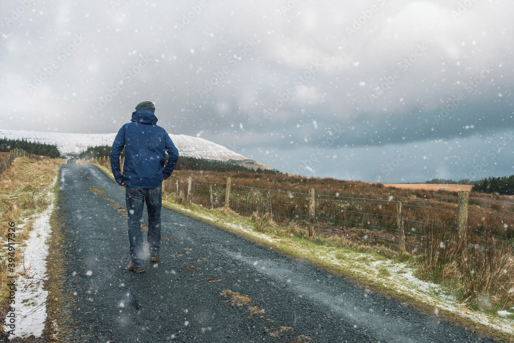 Man in winter clothes walking on a narrow small road to a mountain during snow fall. Winter season concept. Sligo, Ireland.