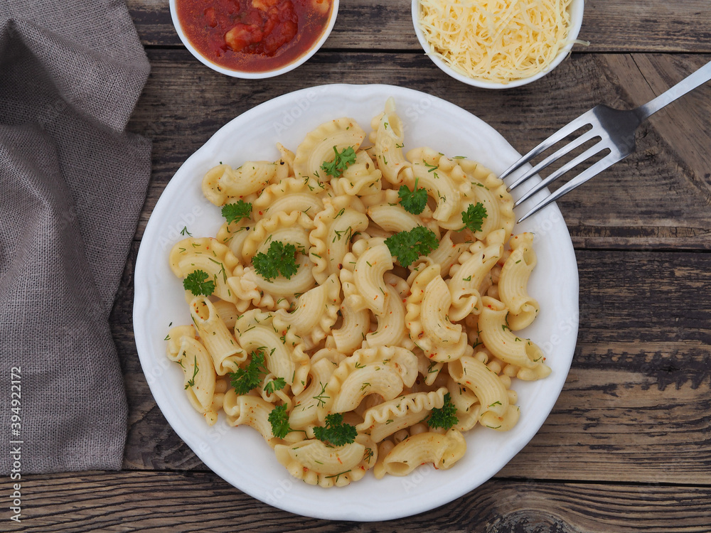 Curly pasta, macaroni with spices and herbs in a plate, tomato sauce, grated parmesan cheese, fork on a wooden table, closeup, top view. Simple delicious rustic dish with pasta products Curly pasta, macaroni with spices and herbs in a plate, tomato sauce, grated parmesan cheese, fork on a wooden table, closeup, top view. Simple delicious rustic dish with pasta products