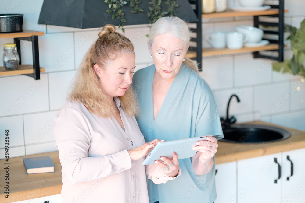 Obraz premium Woman teaching senior mother to use internet at home. Senior woman with her daughter looking at modern gadget indoors. relaxing together, different generations hobby pastime. support for the elderly