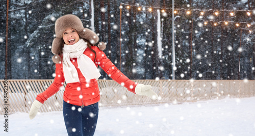 christmas, season and holidays concept - happy woman in winter fur hat having fun over outdoor ice skating rink on background