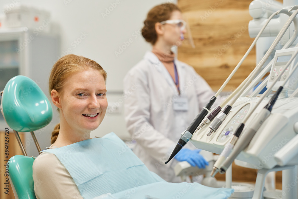 Portrait of young patient smiling at camera while sitting on dental chair with dentist in the background Portrait of young patient smiling at camera while sitting on dental chair with dentist in the background