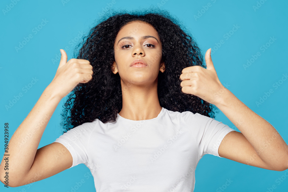Woman with black curly hair white t-shirt showing thumbs up blue background Woman with black curly hair white t-shirt showing thumbs up blue background