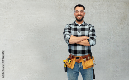 profession, construction and building concept - happy smiling male worker or builder in goggles with crossed arms over grey concrete background