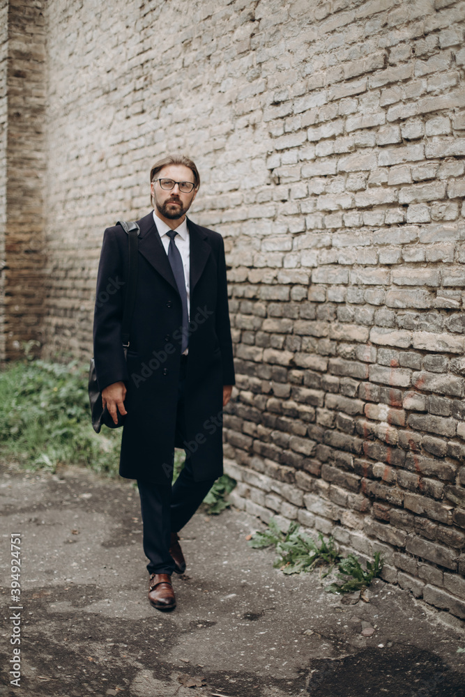 Mature bearded man in formal wear walking near dark brick wall and carrying bag on shoulder. Businessman in eyeglasses going to working meeting. Mature bearded man in formal wear walking near dark brick wall and carrying bag on shoulder. Businessman in eyeglasses going to working meeting.