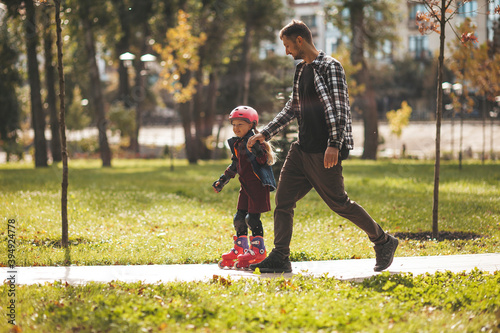 Child girl holding dad's hand, roller skating in the autumn park.