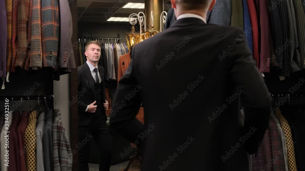 shooting of a man standing in front of the mirror, straightening his sleeves and buttoning the jacket in a luxury men clothing store, against the background of mannequins..