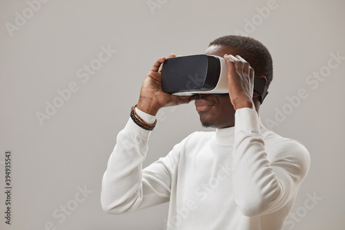 African young man in virtual glasses playing in virtual reality game against the grey background