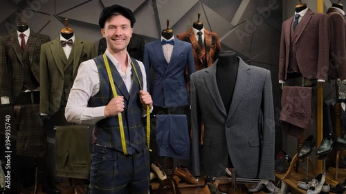 Positive tailor posing in atelier next to mannequin with elegant Blazer or jacket. Portrait of Caucasian man dressed in a vest and a hat, looking at camera and smiling. Male designer in sewing worksho photo