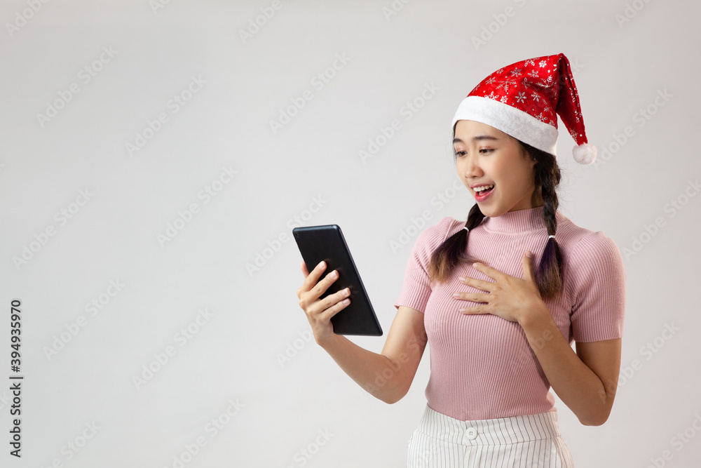 A cute Asian woman wearing a santa hat makes a surprised expression, holding a tablet on a white background A cute Asian woman wearing a santa hat makes a surprised expression, holding a tablet on a white background